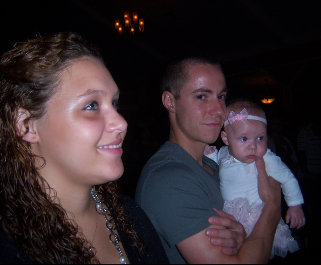 A young woman and young man holding his baby daughter in church. A dark background. The baby has a white headband with a pink bow on it.